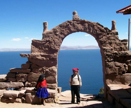 two children standing next to a stne arch with lake titicaca in the background